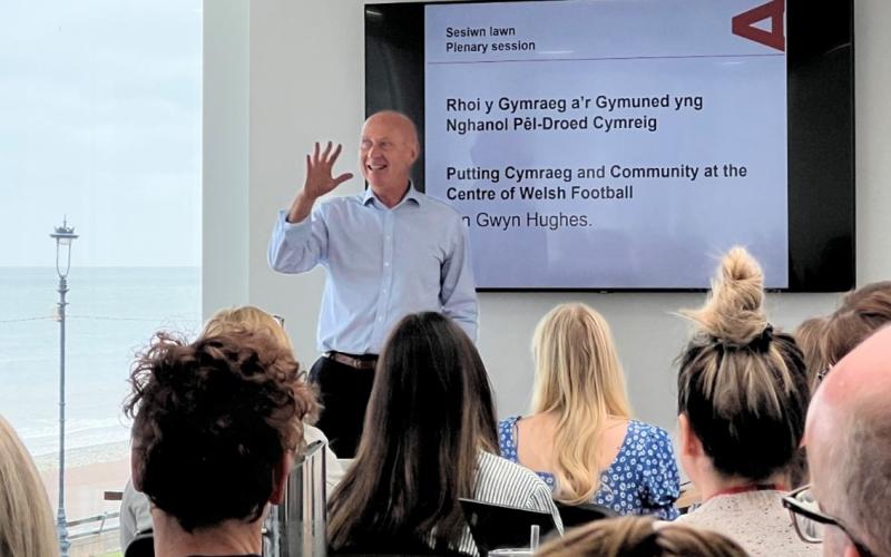 Ian Gwyn Hughes, Pennaeth Cyfathrebu Cymdeithas Bêl-droed Cymru fel y prif siaradwr  ar y llwyfan. Testun y tu ôl iddo yn dweud Rhoi y Gymraeg a'r Gymuned yng Nghanol Pêl-droed Cymreig