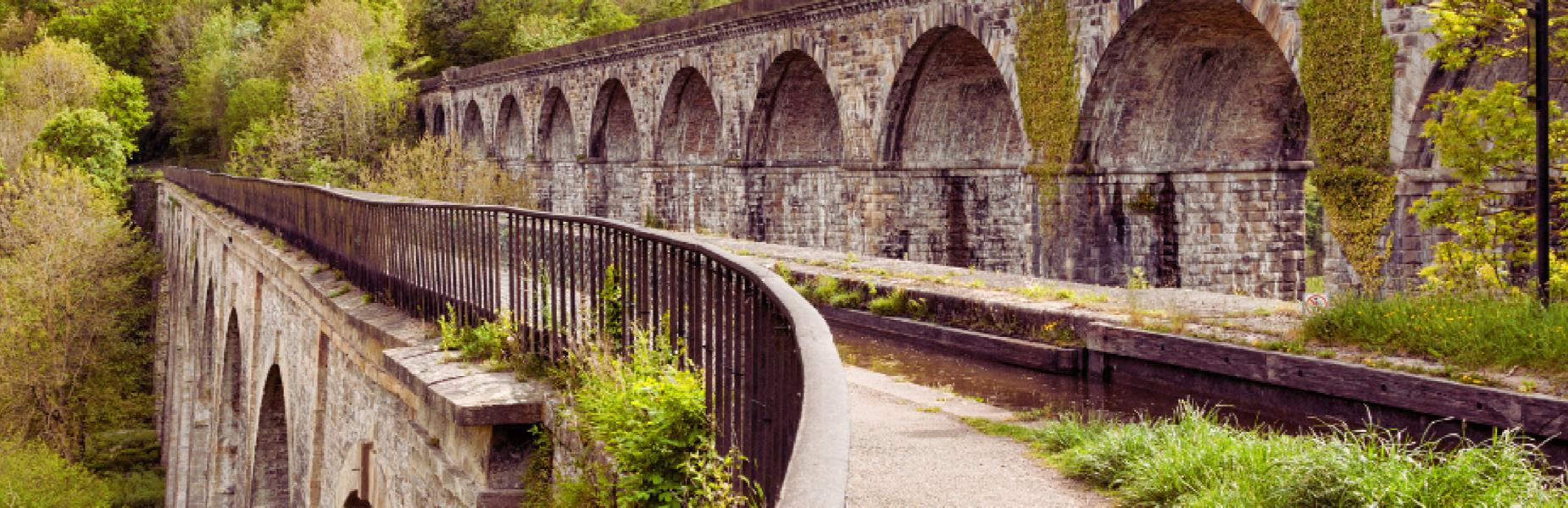 Stone viaduct with arches in Wrexham, surrounded by greenery and a path with railings.